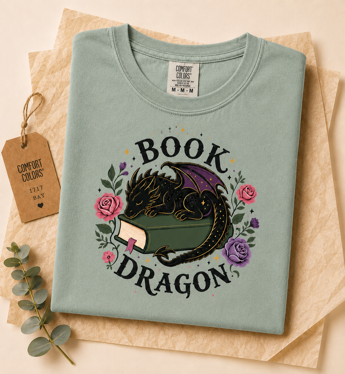 A folded sage green Comfort Colors t-shirt with a 'Book Dragon' graphic, styled on a beige background with brown paper, a decorative eucalyptus sprig, and a paper tag.