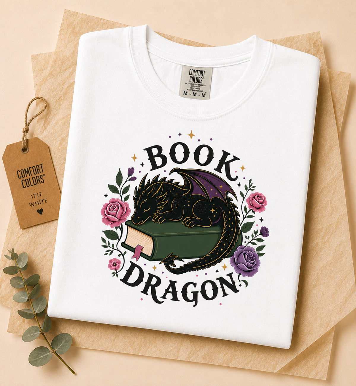 A white Comfort Colors t-shirt laid flat on brown kraft paper, featuring a graphic of a black dragon curled over an open green book, surrounded by pink and purple roses and the words 'BOOK DRAGON'. A matching paper tag and eucalyptus sprig are placed beside the shirt.