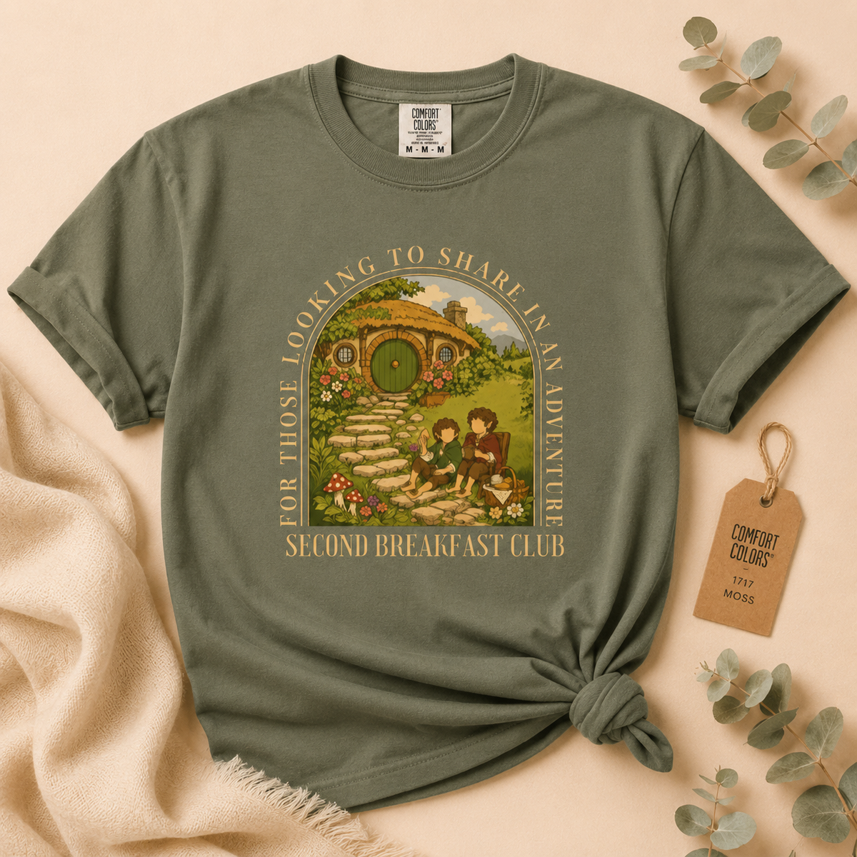 An olive green graphic t-shirt styled in a flat-lay arrangement with a beige blanket, eucalyptus leaves, and a paper hangtag. The t-shirt features a colorful graphic of hobbits at a breakfast table in front of a hobbit house, with the text 'FOR THOSE LOOKING TO SHARE IN AN ADVENTURE' and 'SECOND BREAKFAST CLUB'.